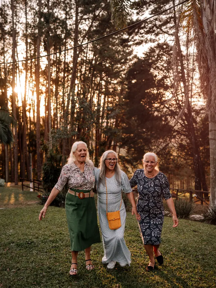 Three older women walking arm in arm through a sunlit forest