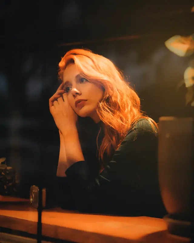 A woman with long red hair resting her chin on her hand in a lamp-lit cafe